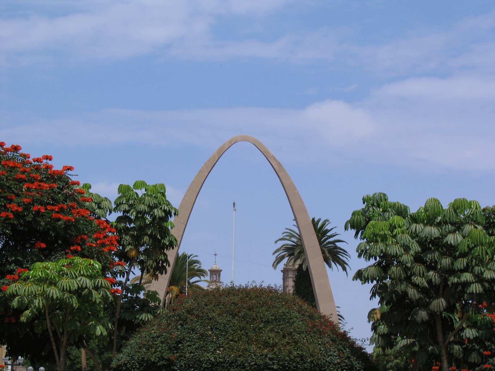 Arco Parabólico de Tacna entre árboles en flor, con las torres de la catedral detrás